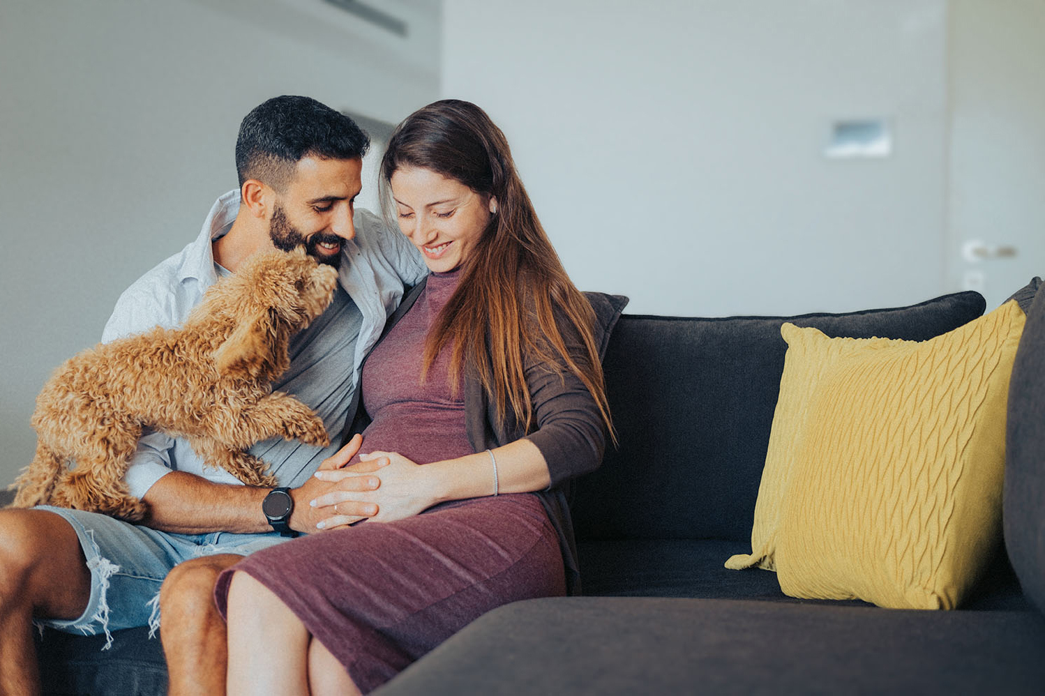 אישה בהריון ובן זוגה יושבים שמחים על הספה ועליהם כלב