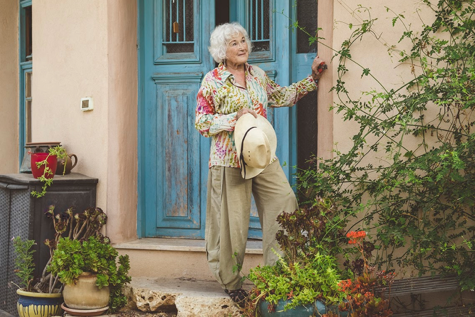 אישה מבוגרת עומדת ומביטה בחצר, כשבידה כובע