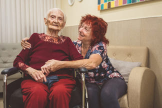 אישה בגיל השלישי יושבת על כיסא גלגלים ומחבקת אותה מהצד אישה שיושבת על ספה