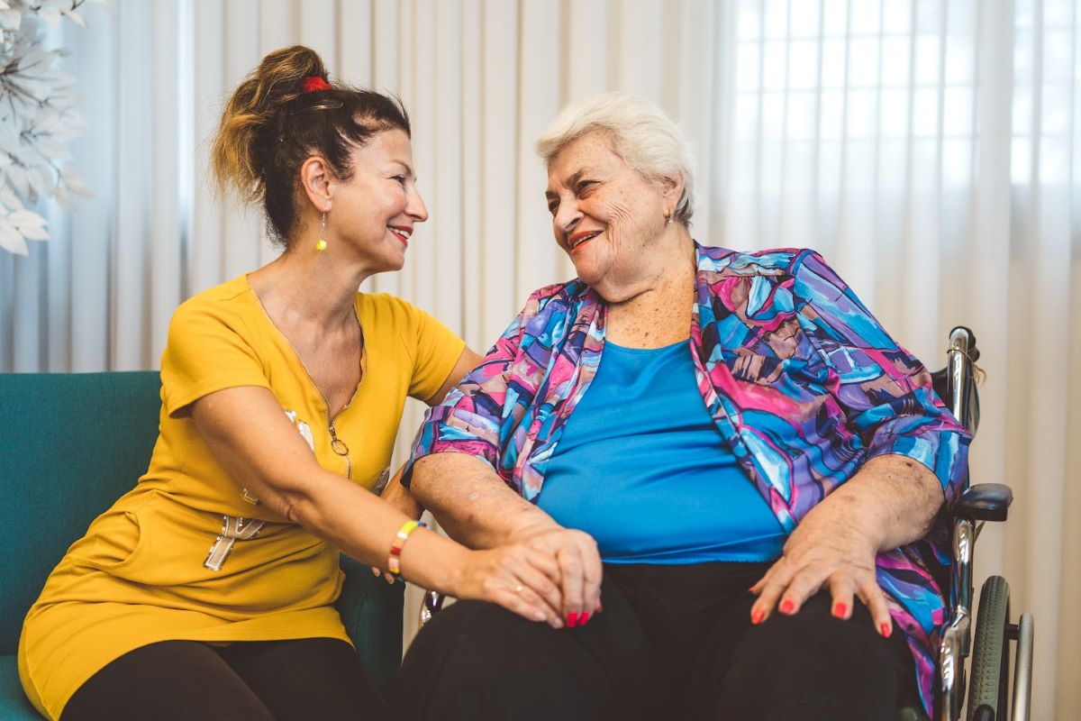 אישה יושבת ומחזיקה יד של אישה בגיל השלישי שיושבת על כיסא גלגלים