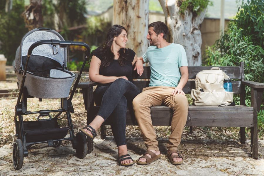 זוג הורים צעירים יושבים על ספסל בגינה, מחייכים אחד לשנייה, כשלידם עגלת תינוק