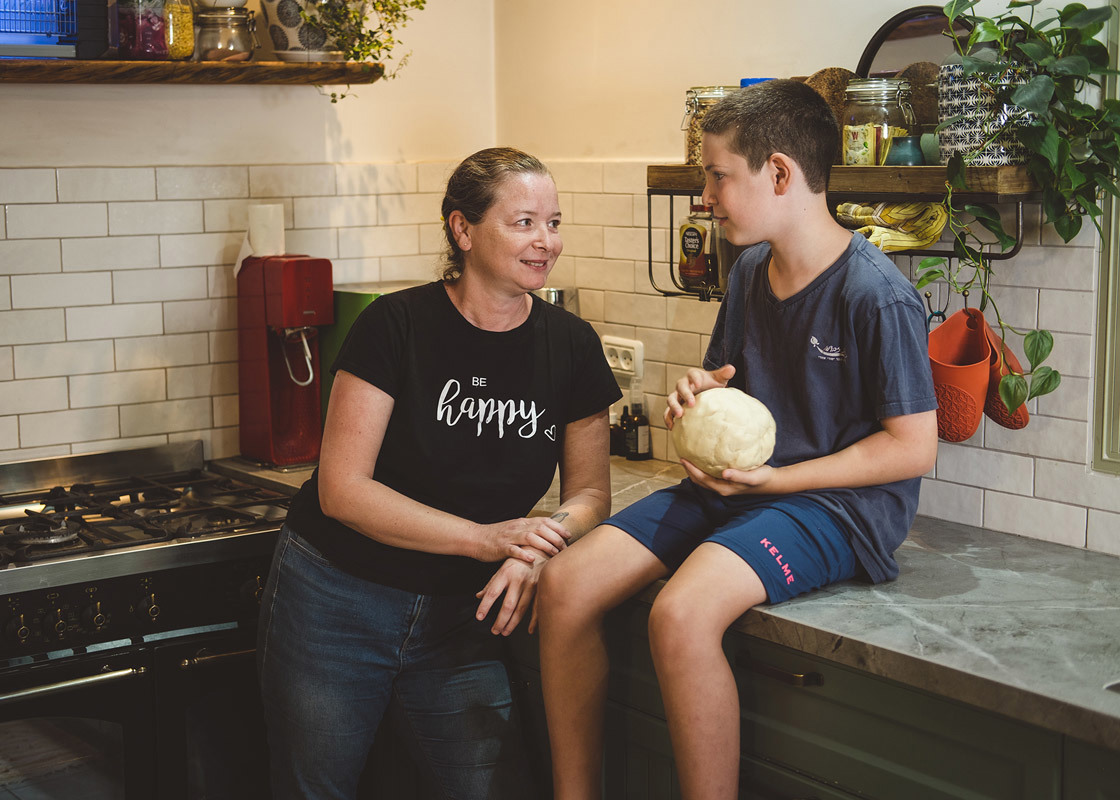 Подросток сидит на кухонной столешнице с шаром теста и разговаривает с женщиной, стоящей рядом с ним