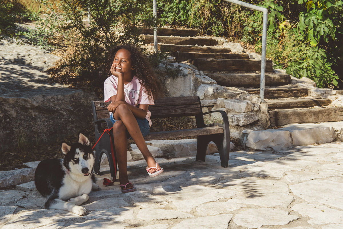 A girl is sitting on a bench, smiling, holding the leash of a Siberian Husky dog sitting beside her.