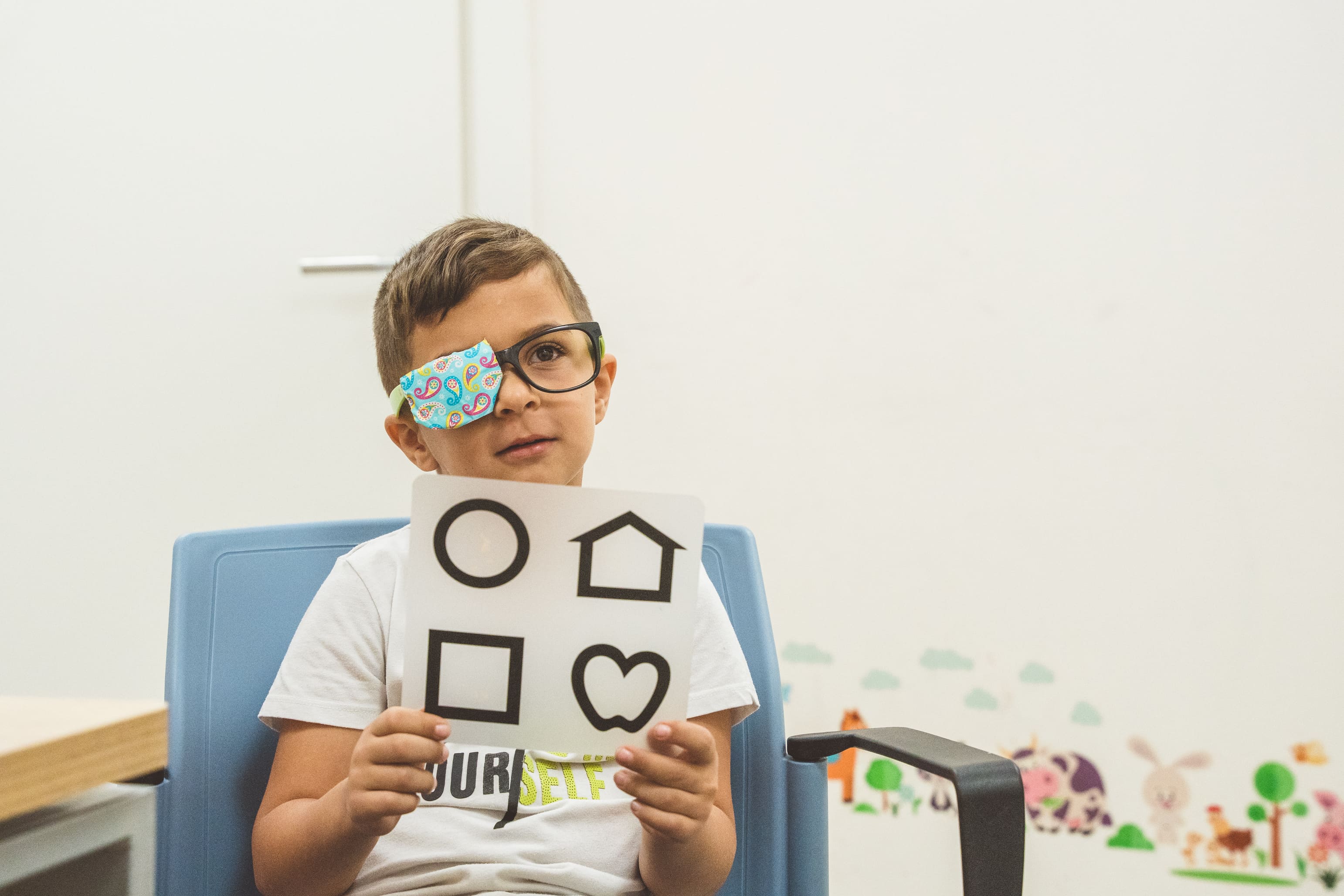 A boy with a bandage on his eye is holding a card with shapes.