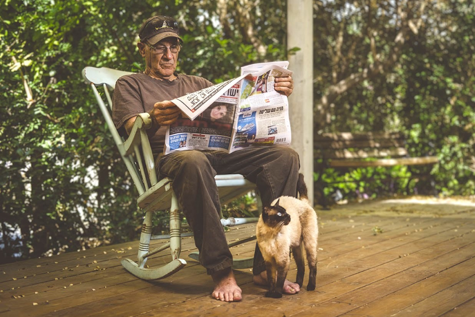 Elderly man sitting on a swing chair in the yard, reading a newspaper, a cat beside him