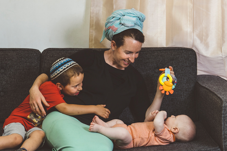 אישה יושבת על ספה חובקת את בנה הגדול ושניהם מבינים בשמחה בתינוק