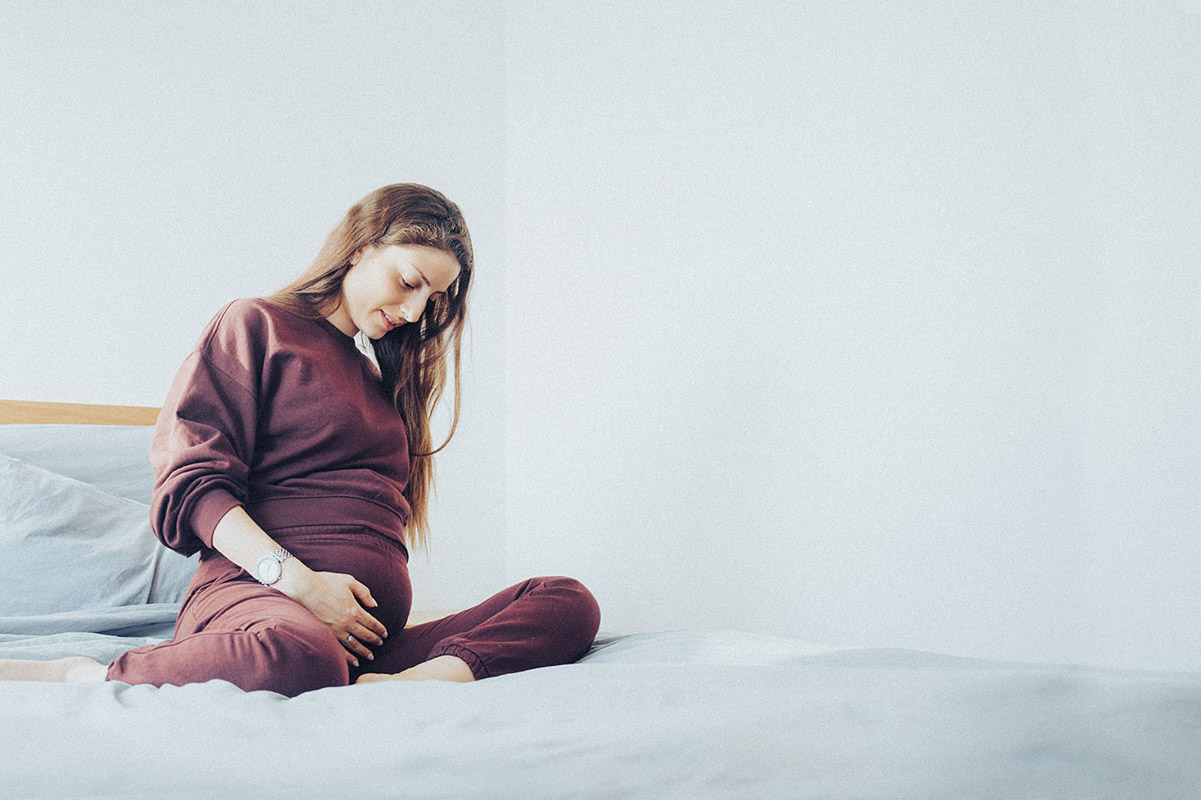 אישה צעירה בהריון יושבת על מיטה, מתבוננת בבטן שלה ומחזיקה בה