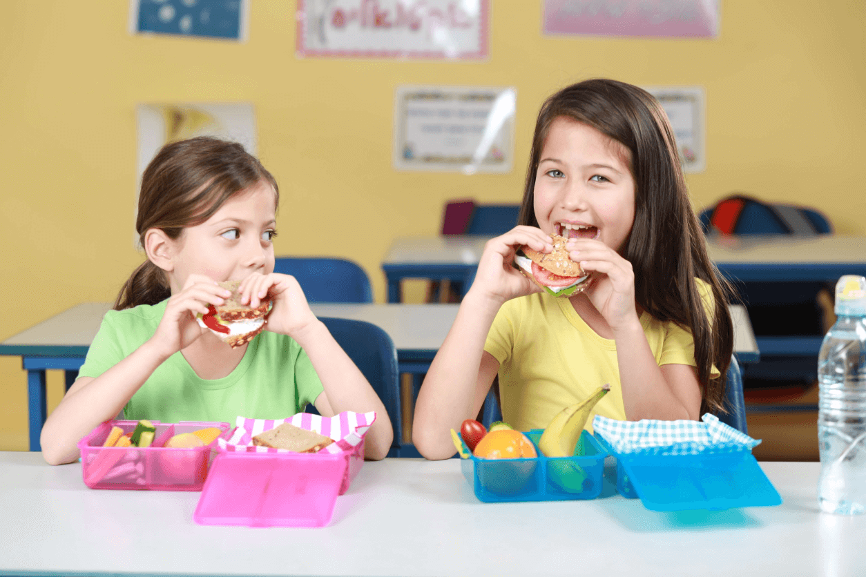 Two girls are eating sandwiches over lunchboxes.