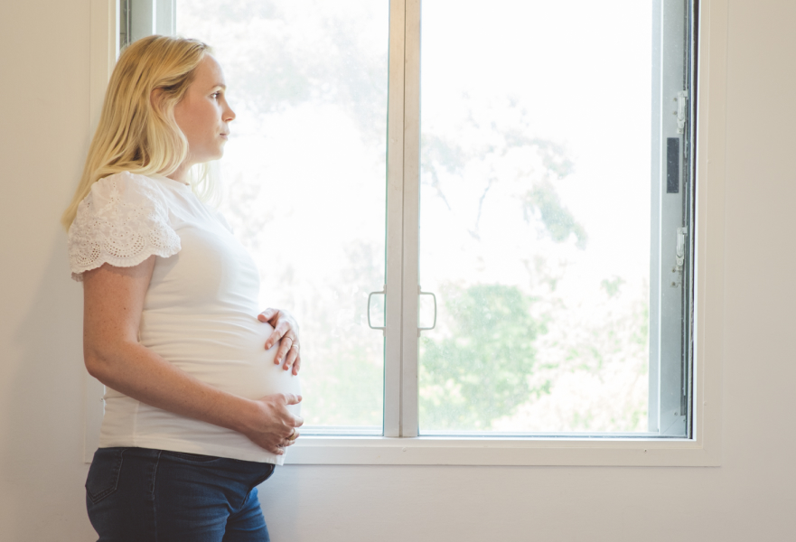 אישה בהריון עומדת ומביטה החוצה מהחלון